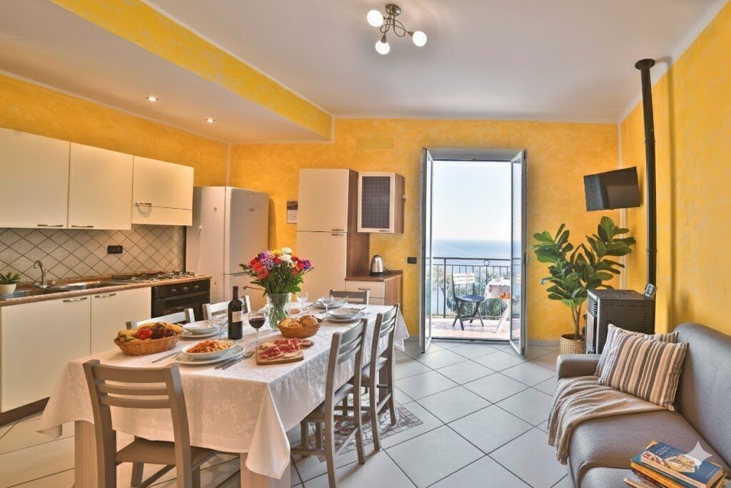 Cozinha luminosa com armários brancos, uma mesa de jantar posta para refeição e uma varanda com vista.