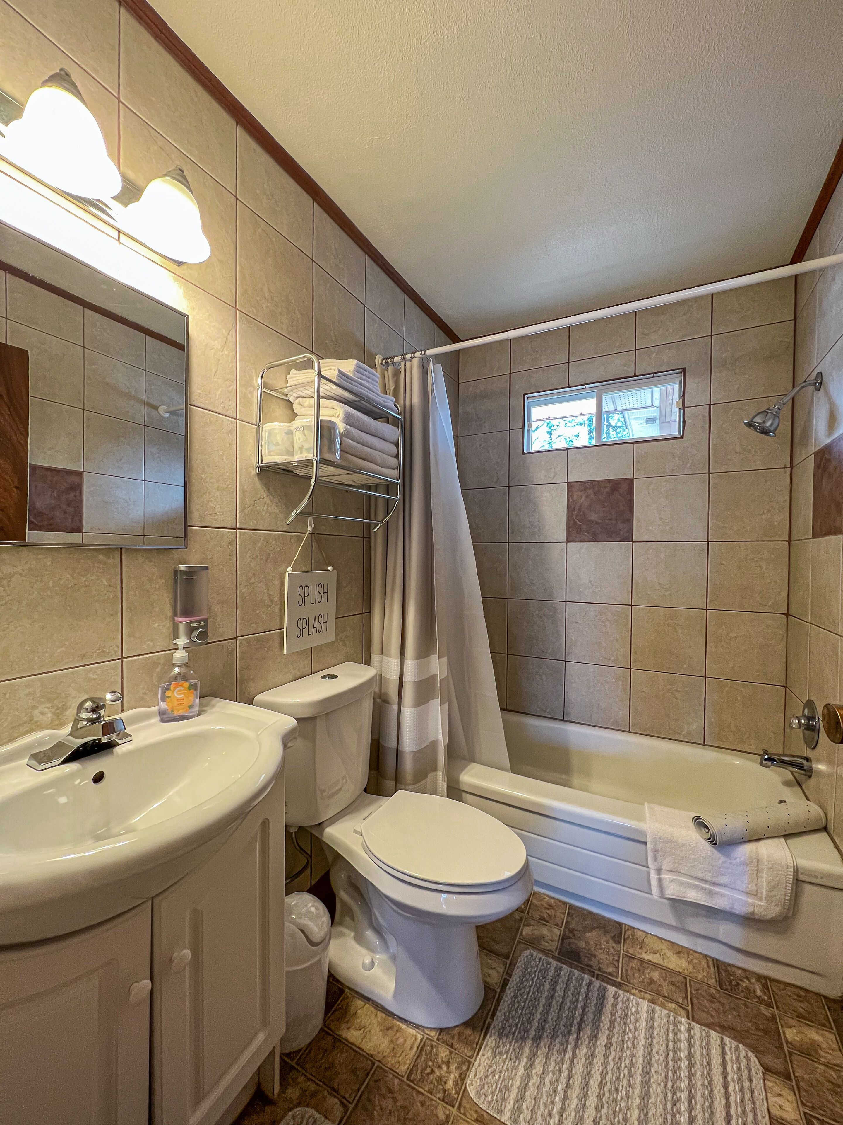 Een badkamer met toilet, wastafel en ligbad. Er is een douchegordijn, een handdoekrek en een raam.
