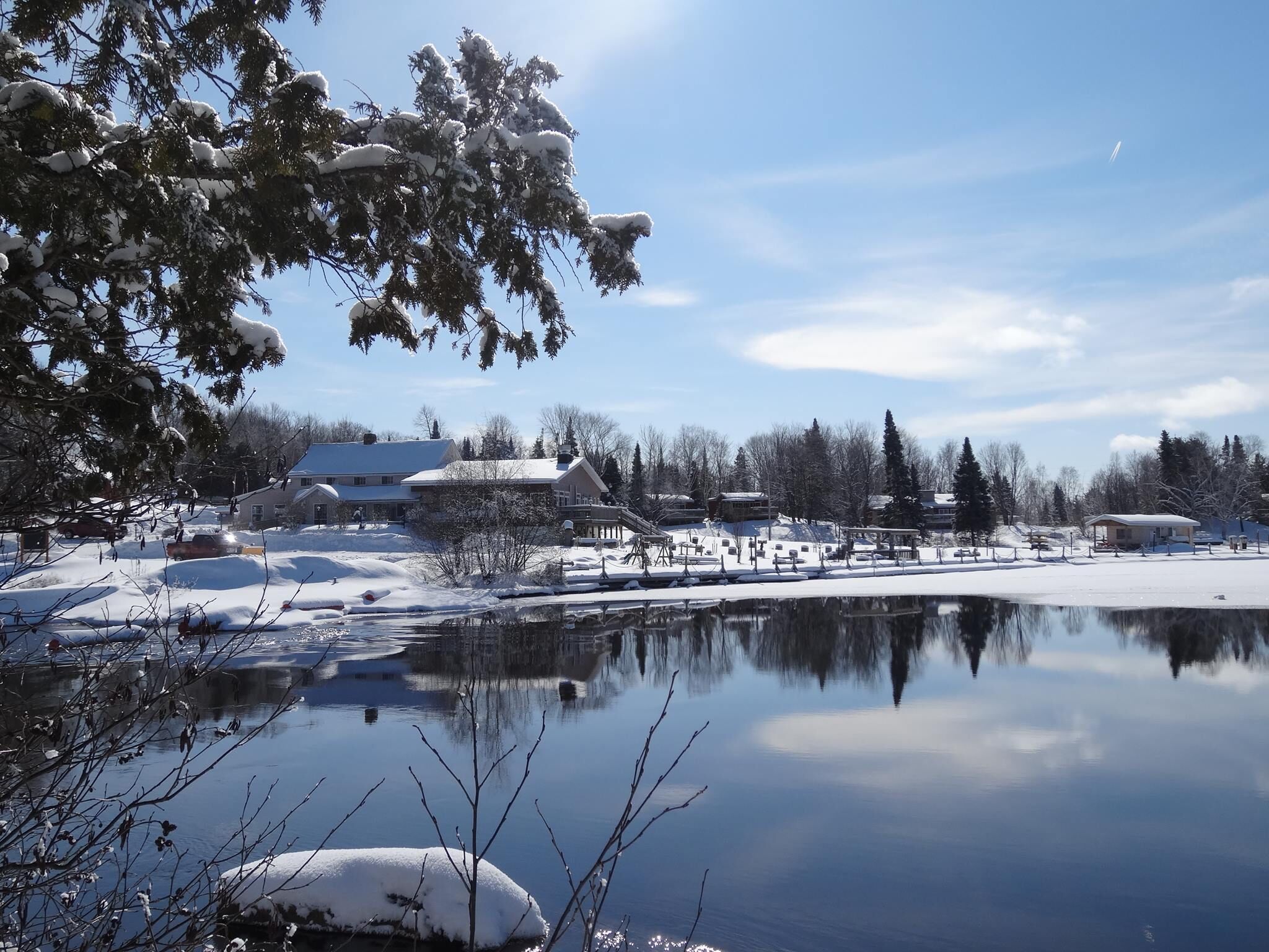 Et fredfyldt vinterlandskab med sø, der spejler bygninger og træer, omgivet af sneklædt jord og klare himmel.