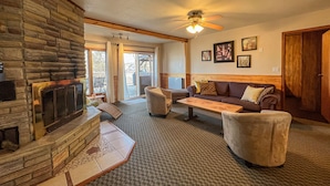 A cozy living room with a stone fireplace, a ceiling fan, and a large window with curtains. - Ahmic Lake Resort's 2-Bedroom Cottage with Hot Tub (Gray Wolf Cottage) (Magnetawan)