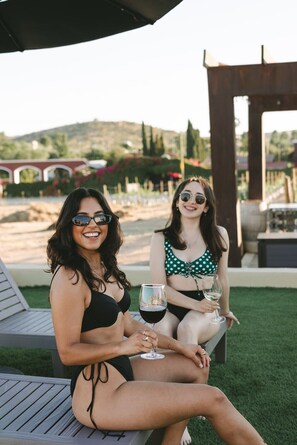 Two people sitting on outdoor lounge chairs with drinks, in a setting with a pergola and a view of hills in the background.