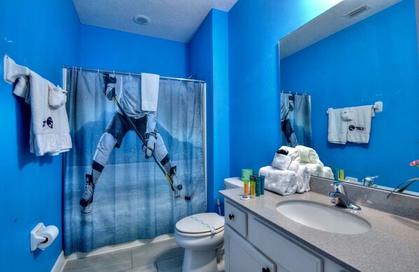 A bathroom with blue walls, a shower curtain with a person climbing a ladder, a toilet, a sink with toiletries, and a mirror. - Bowling, Sports-themed, Private Pool, 1GB Wi Fi, video wall TV, sleeps 40 (Kissimmee)