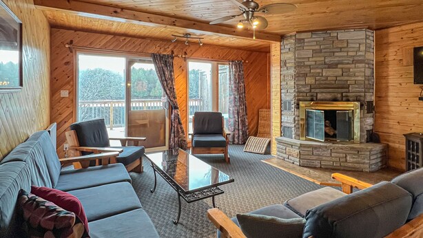 A cozy cabin interior with a stone fireplace, wooden furniture, and large windows. - Ahmic Lake Resort's 3-Bedroom Cottage (Red Fox Cottage) (Magnetawan)