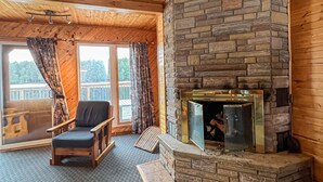 A cozy cabin interior with a stone fireplace, wooden ceiling, and a comfortable chair by a large window. - Ahmic Lake Resort's 3-Bedroom Cottage (Red Fox Cottage) (Magnetawan)