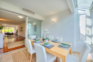 Ein Essbereich mit einem Holztisch, gedeckt für sechs Personen, weißen Stühlen und einer Glastür, die in einen Wohnbereich führt.
