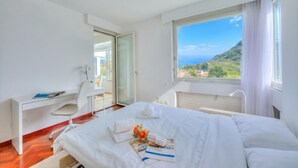 A hotel room with a bed, a desk, a chair, a window with a view of a mountain and sea, and a bathroom door.
