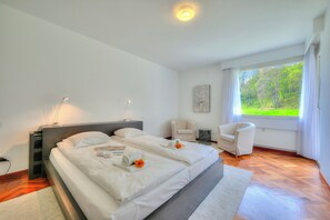 Ein modernes Schlafzimmer mit einem großen Bett, Nachttischlampen, einem Sessel und Blick auf Grünfläche.