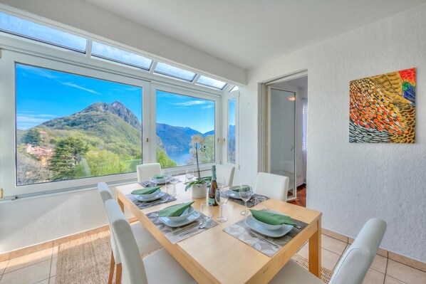 Un coin repas avec une table dressée pour quatre personnes, de grandes fenêtres offrant une vue sur les montagnes et un tableau abstrait aux couleurs vives accroché au mur.