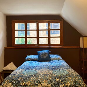 Une chambre avec une grande fenêtre, un lit recouvert d’une literie à motifs et une table de chevet avec un oreiller.