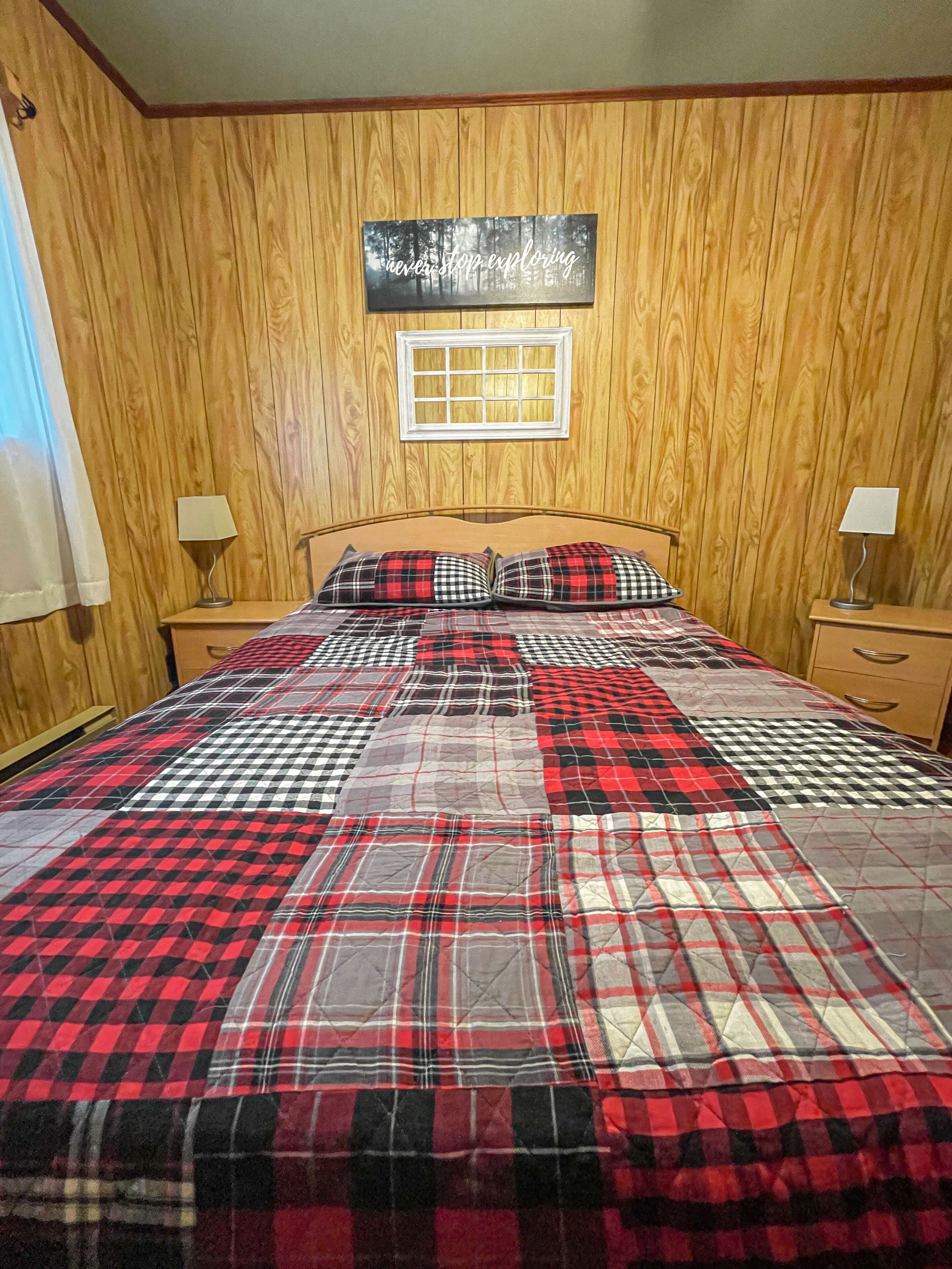 Une chambre avec un mur lambrissé en bois, un lit recouvert d’une couette à carreaux rouges et noirs, deux tables de chevet avec des lampes, et une fenêtre donnant sur des arbres.