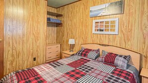 Un dormitorio con pared de paneles de madera, una cama con colcha escocesa, un ventilador de techo, una mesita de noche con lámpara y un cuadro en la pared.