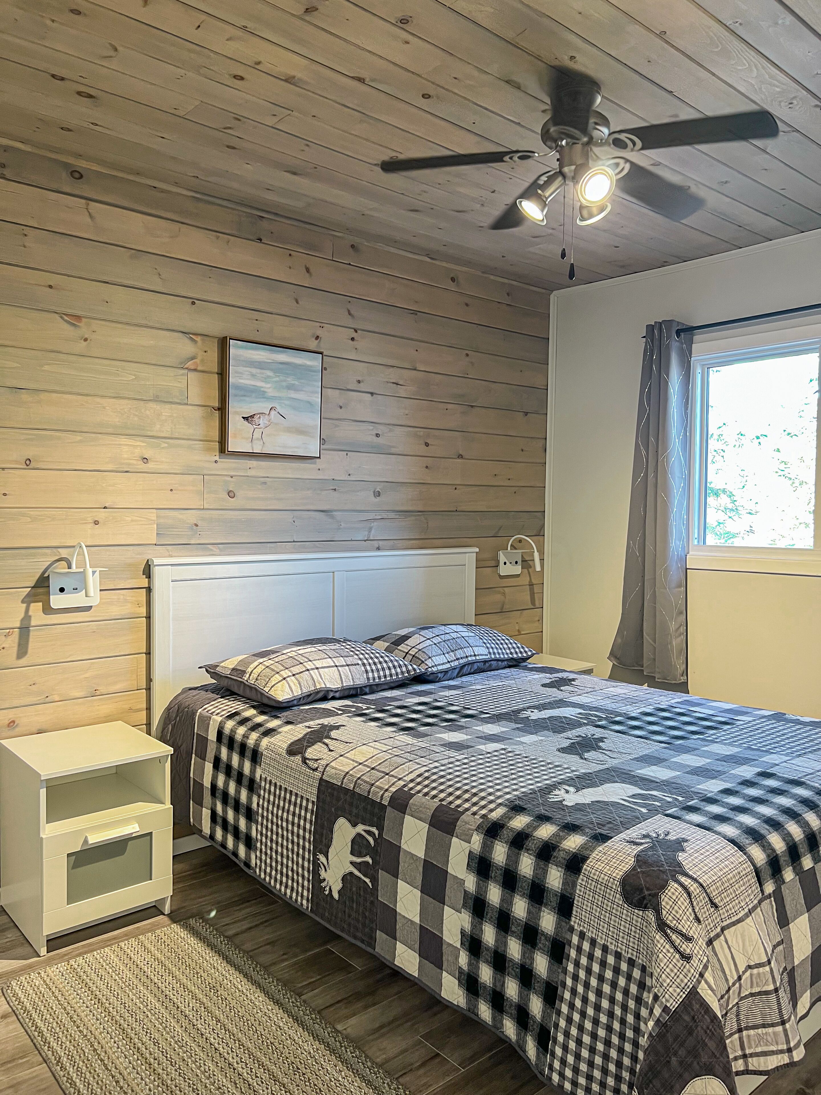 A bedroom with a wooden ceiling, a bed with a checkered bedspread, a nightstand, a bedside table, a ceiling fan, and a window with curtains.