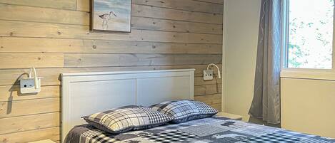 A bedroom with a wooden ceiling, a bed with a checkered bedspread, a nightstand, a bedside table, a ceiling fan, and a window with curtains.