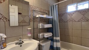Un baño con lavabo, inodoro y bañera. Hay un estante con toallas y una ventana sobre la bañera.