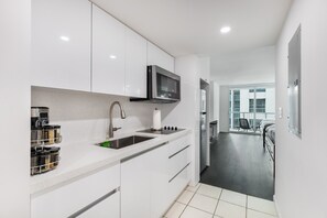A modern kitchen with white cabinets, a stainless steel sink, and a microwave. There is a small dining area with a table and chairs. - Lovely Brickell Oceanview Studio (Miami)