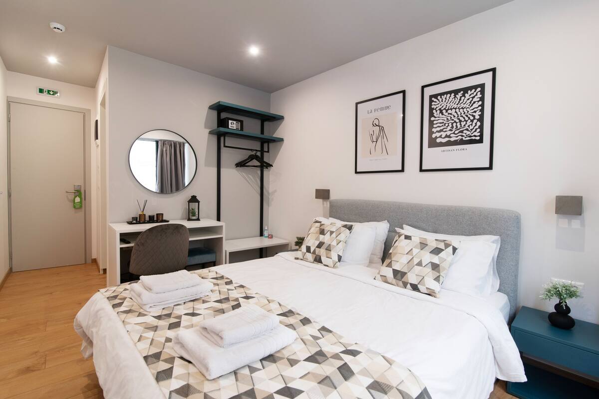 Una habitación de hotel con cama, escritorio, silla, espejo y obras de arte en las paredes.