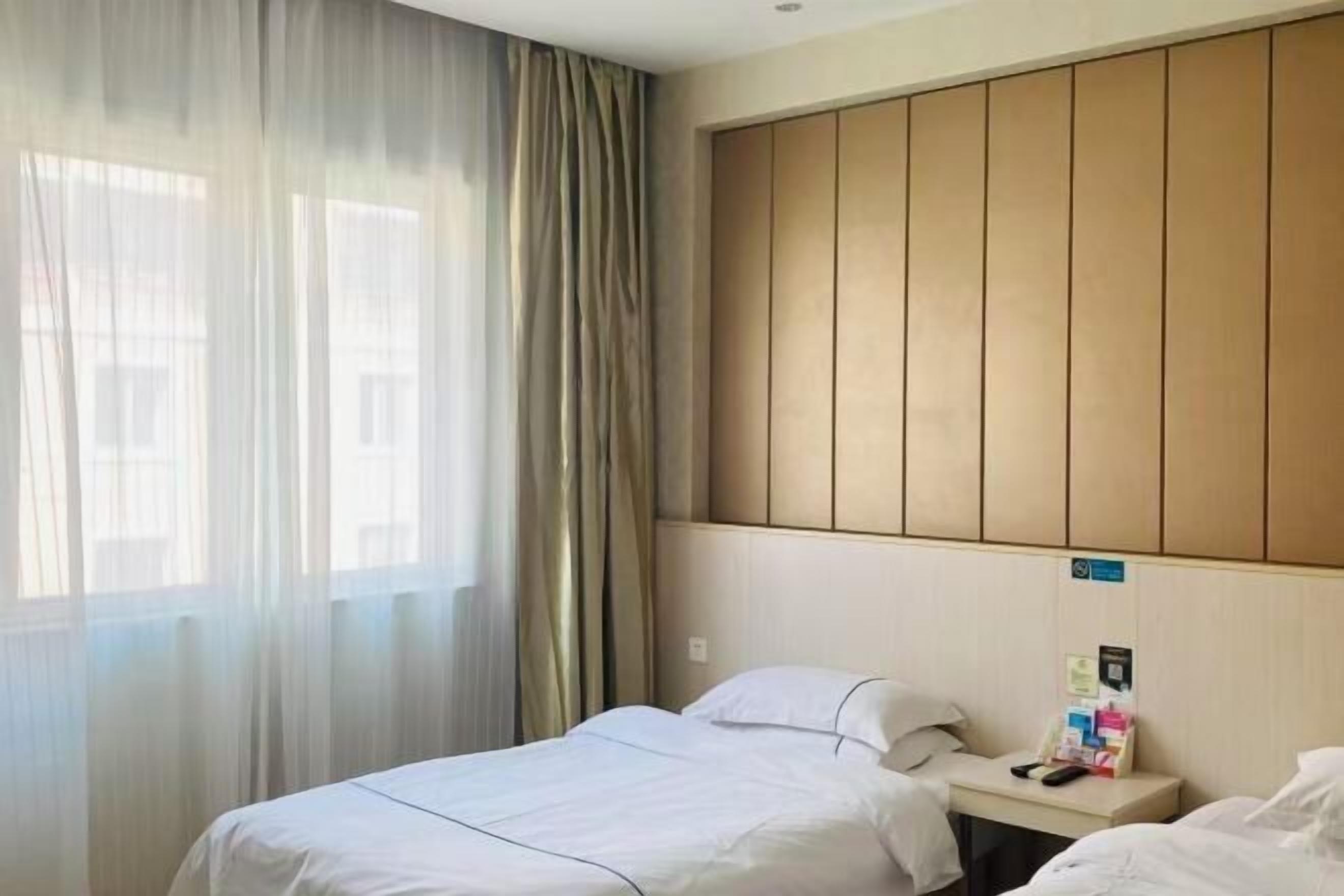 Habitació d'hotel amb un llit, una tauleta de nit, una finestra amb cortines transparents i un panell de fusta al capçal.