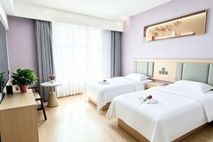 Quarto de hotel com duas camas, uma escrivaninha, uma cadeira, uma mesinha e uma planta em vaso.