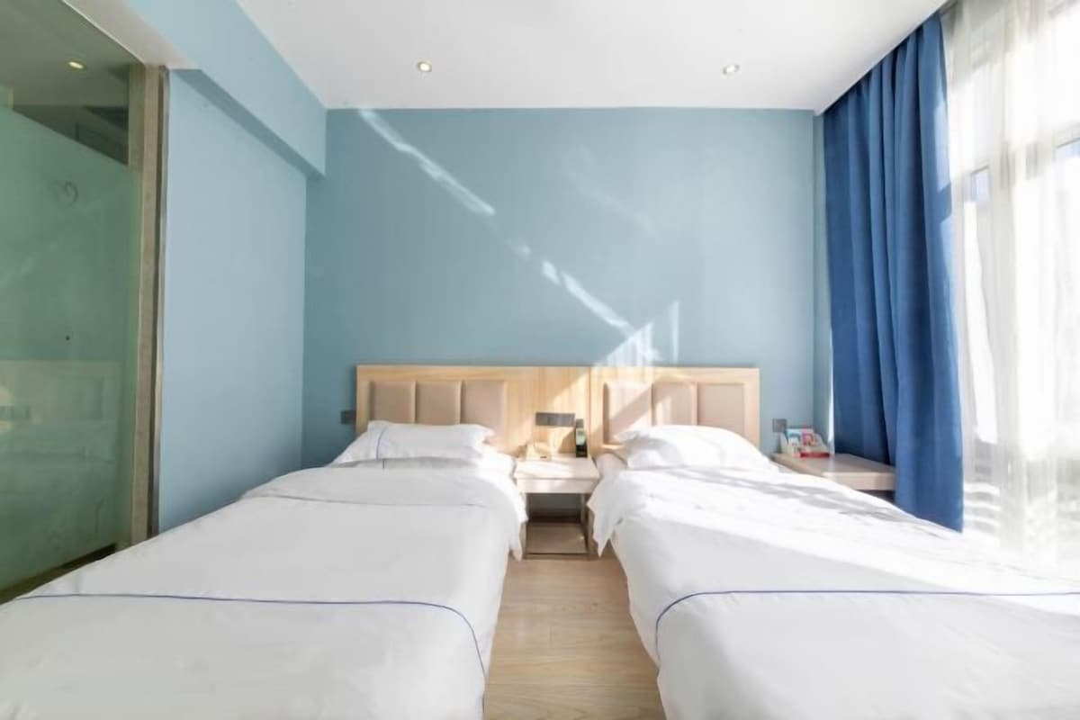 Habitación de hotel con dos camas, un cabecero de madera, una mesita de noche con lámpara y una ventana con cortinas azules.