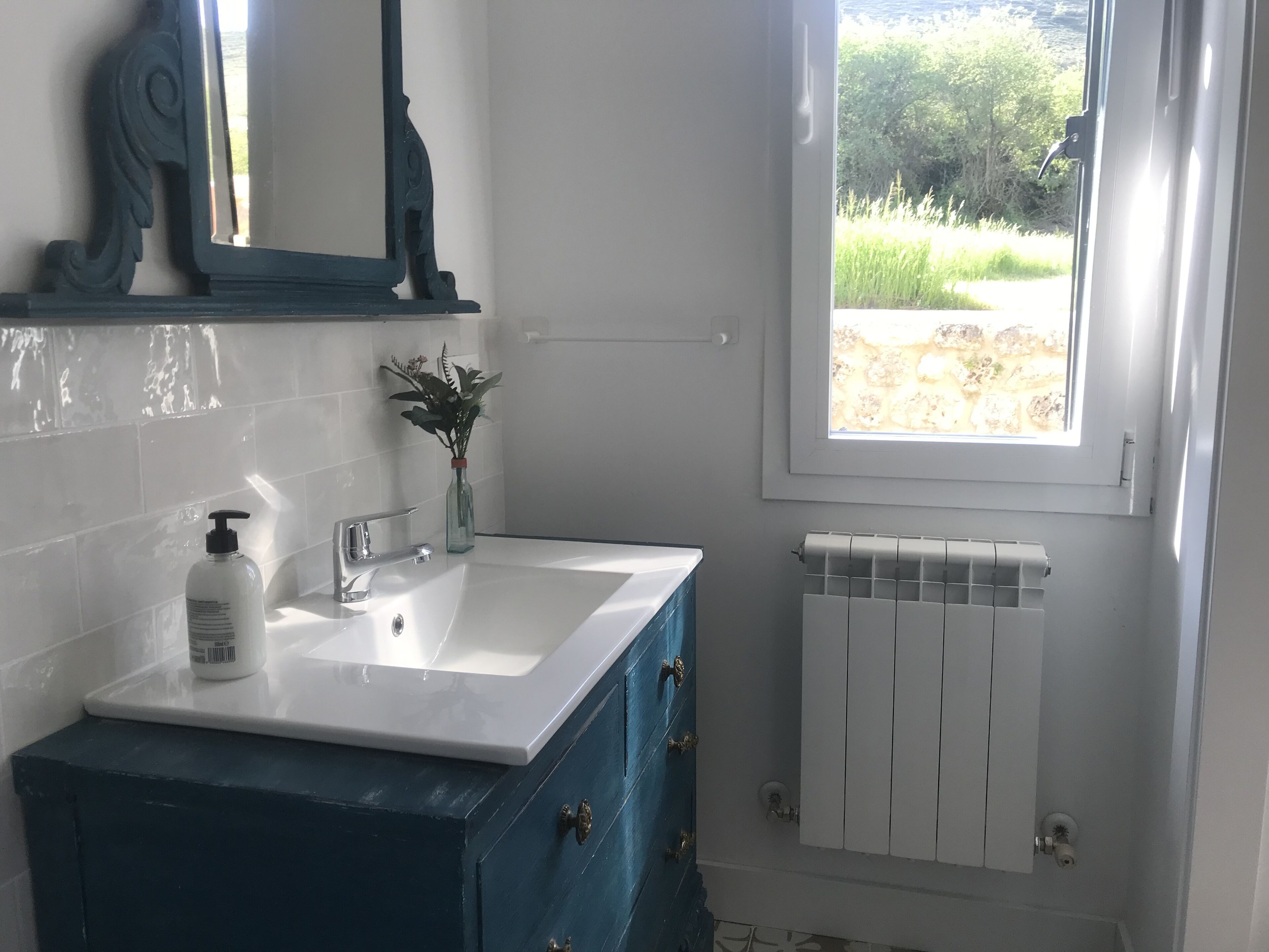 A bathroom with a white sink, a blue cabinet, a mirror, a window, and a radiator.