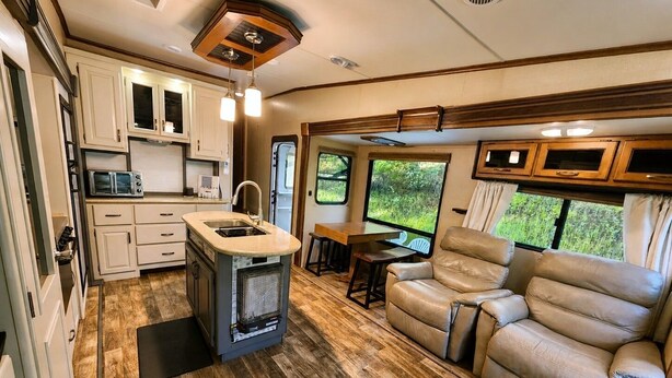 A compact RV interior with a kitchenette, a seating area, and a large window offering a view of greenery.