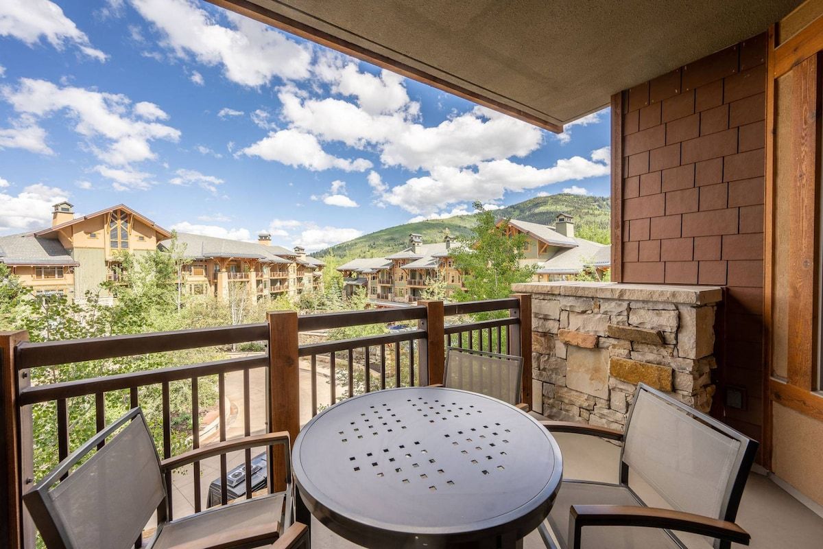 A balcony with a round table and chairs, overlooking a mountainous landscape with multiple buildings.
