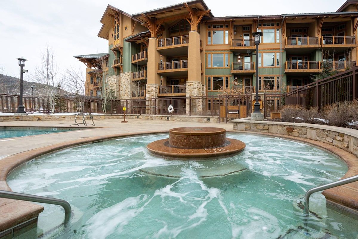 Un área de spa con un jacuzzi rodeado de paredes de piedra, una terraza de madera y un edificio con varios balcones al fondo.