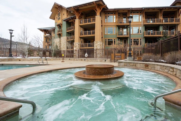 A spa area with a hot tub surrounded by stone walls, a wooden deck, and a building with multiple balconies in the background.