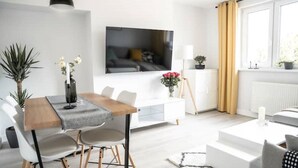 Ein modernes Wohnzimmer mit einem Holzesstisch, weißen Sofas, einem an der Wand montierten Fernseher und einem großen Fenster mit Vorhängen.
