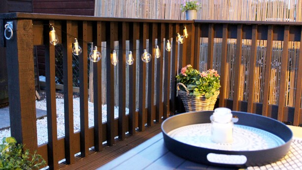 Eine Holzterrasse mit einem runden Tisch, einem weißen Gegenstand auf dem Tisch und Lichterketten.