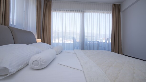 A hotel room with a large bed, a view of the city through sheer curtains, and a balcony with a table and chairs. - Helena Apartments (Budva)