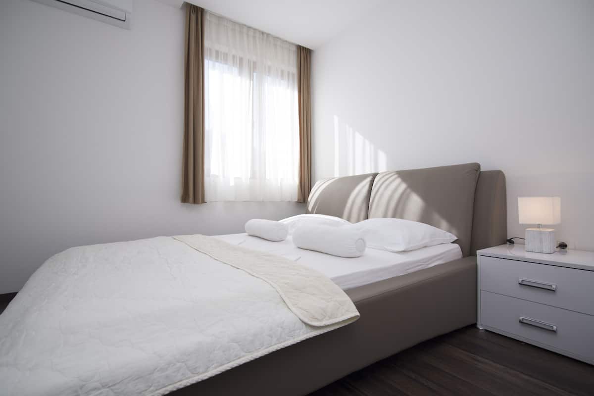 Ein ordentlich bezogenes Bett mit weißer Tagesdecke, einer Nachttischlampe und einem Nachttisch mit Schubladen.