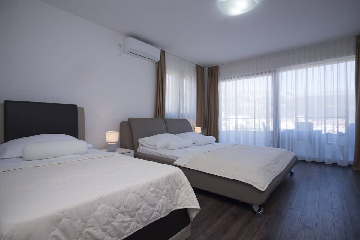 Ein Hotelzimmer mit zwei Betten, einem großen Fenster mit transparenten Vorhängen, einem Deckenventilator und einer Klimaanlage.