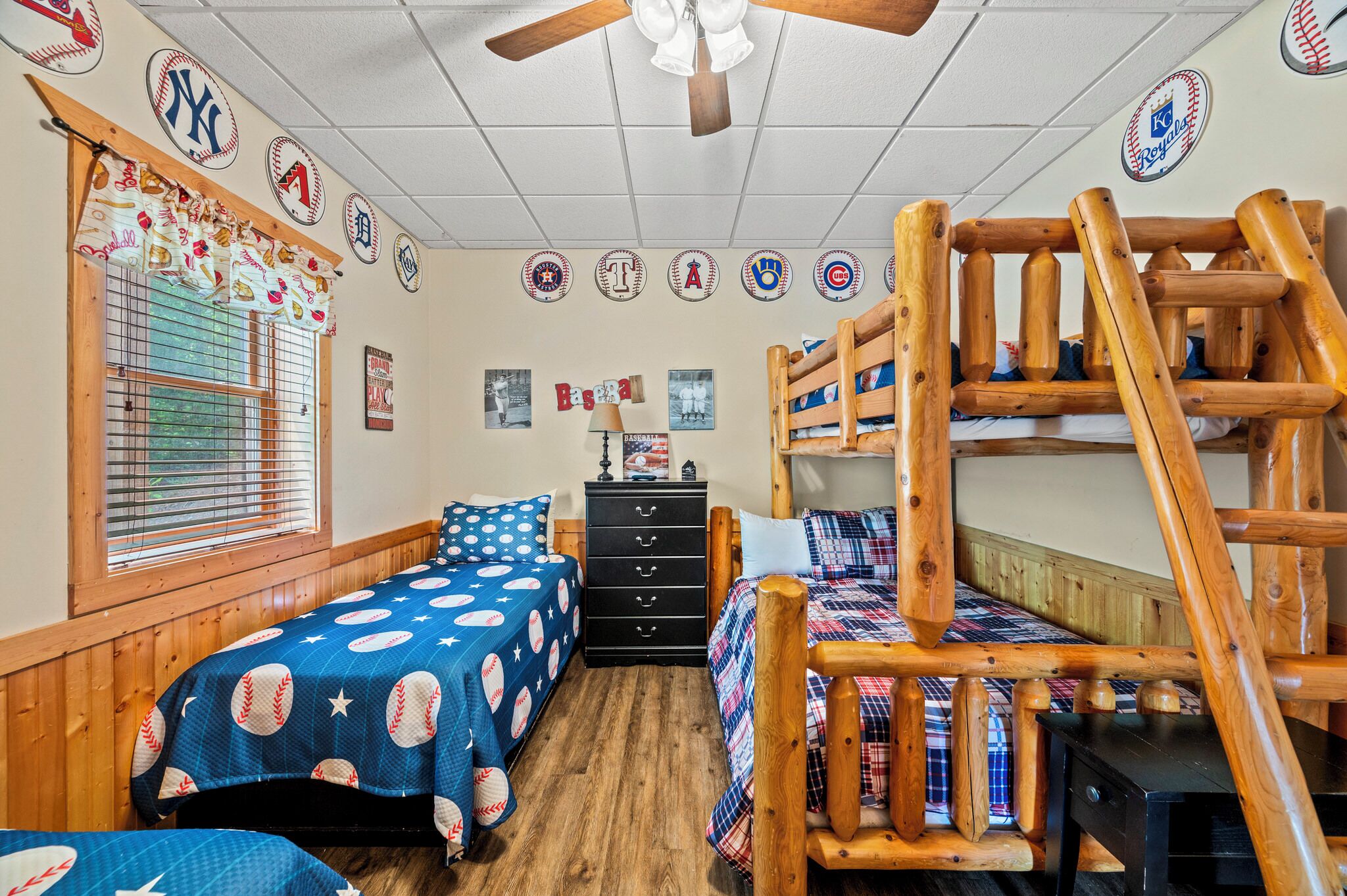 A room with two beds, a wooden bunk bed, a window with blinds, and baseball-themed decor.