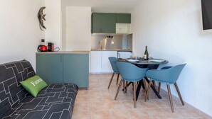 Un espace de vie compact comprenant un coin cuisine, un coin repas avec quatre chaises, un canapé recouvert d’un tissu à motifs et un coussin vert.