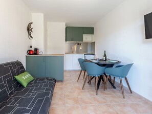 Un espace de vie compact comprenant un coin cuisine, un coin repas avec quatre chaises, un canapé recouvert d’un tissu à motifs et un coussin vert.