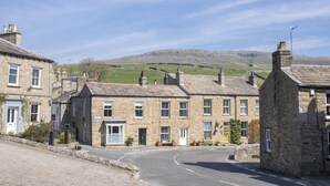 A stone building with a traditional design, featuring multiple windows and doors, situated in a rural setting with a clear sky and distant hills. - Brook House, Askrigg, Yorkshire Dales (Askrigg,)