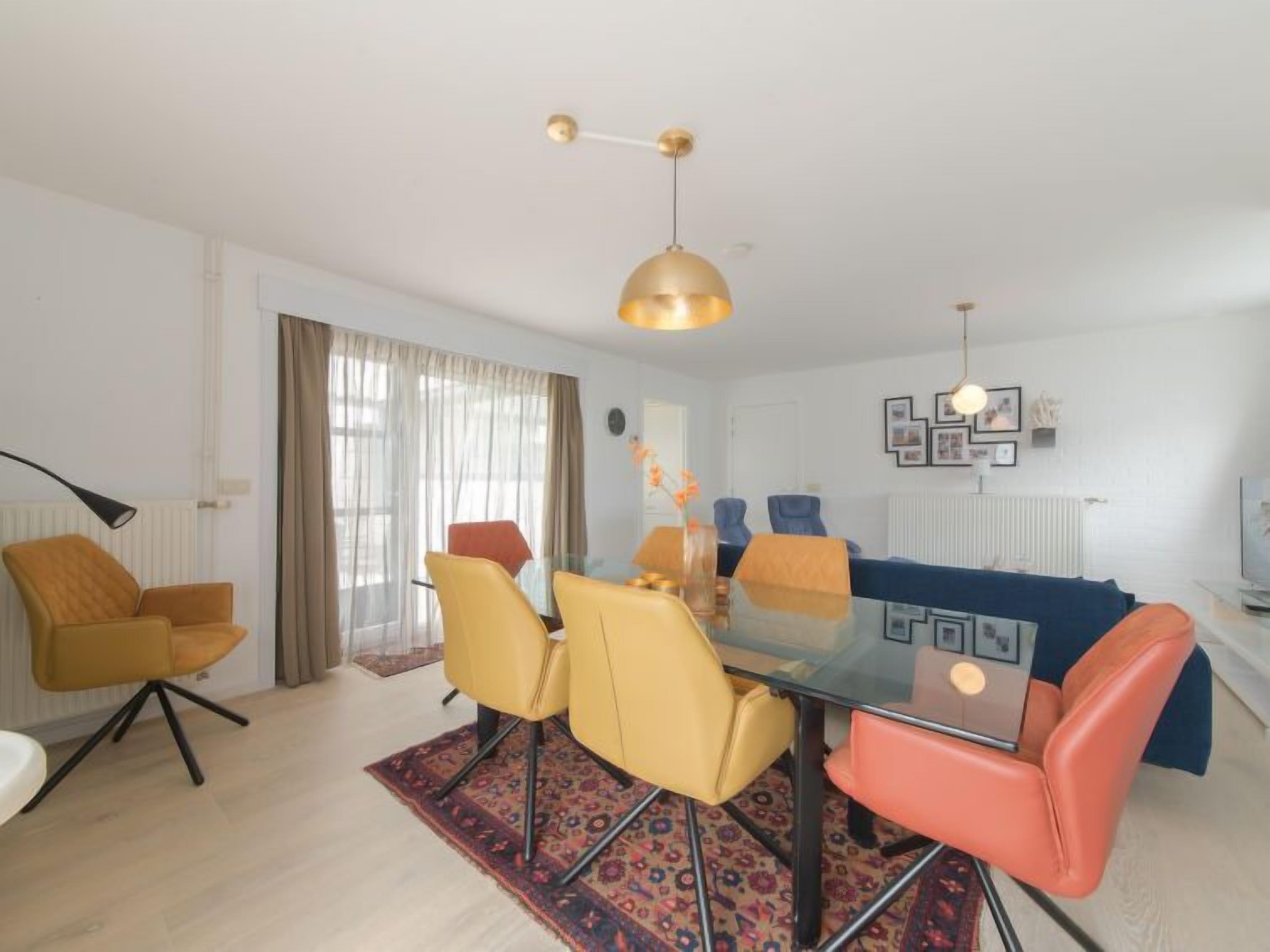 A modern living room with a glass dining table, yellow and orange chairs, a blue sofa, and a patterned rug.