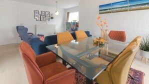 A modern living room with a glass-top dining table, blue and orange chairs, a large window, and a beach painting on the wall. - Klemskerkestraat with terrace by Interhome (Bredene)