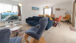 A modern living room with a blue sofa, a dining area with orange chairs, and a flat-screen TV on a glass table. - Klemskerkestraat with terrace by Interhome (Bredene)