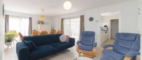 A modern living room with a blue sofa, two blue armchairs, and a white coffee table. There is a dining area in the background with a table and chairs.