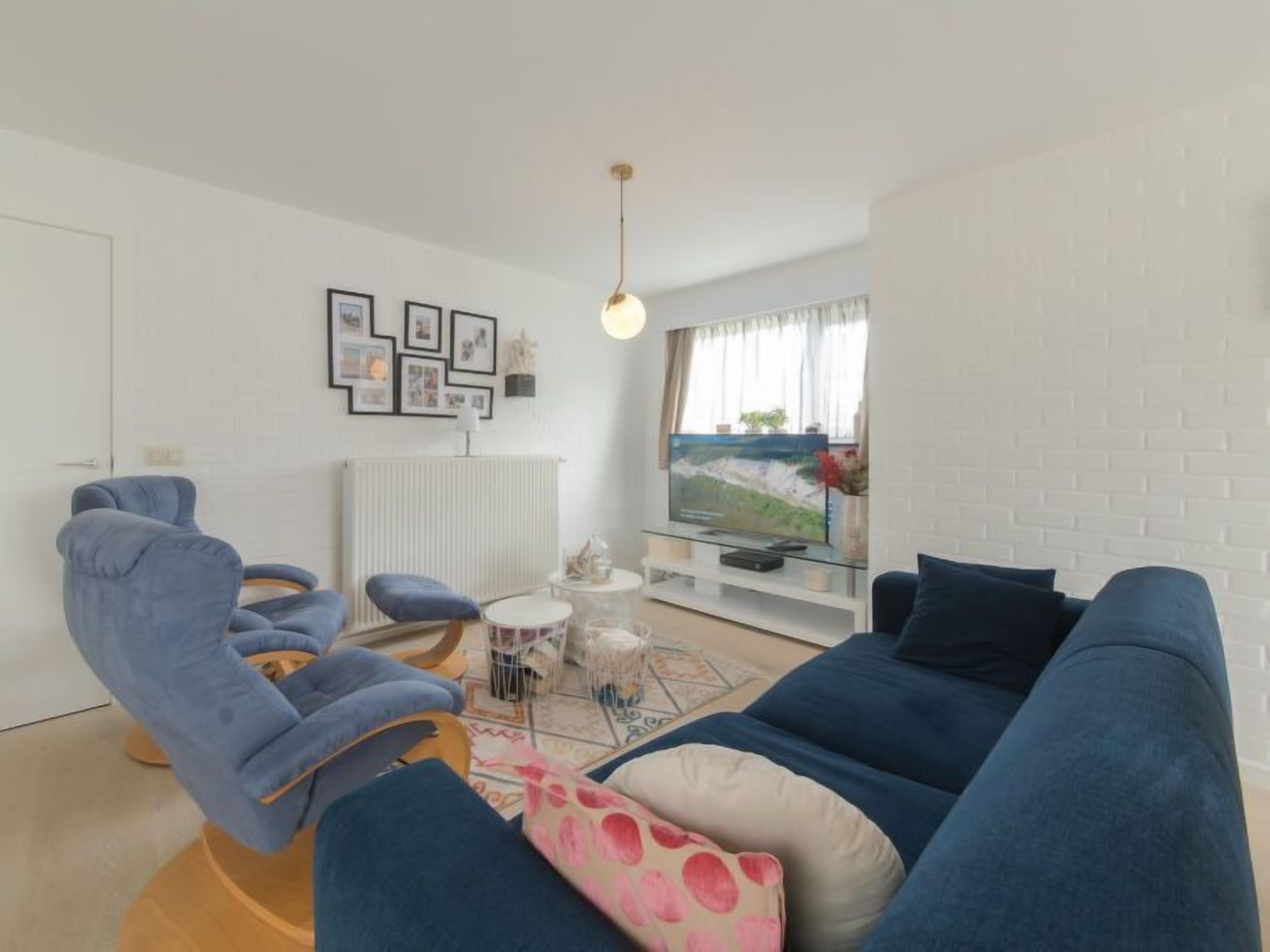 A modern living room with a blue sofa, a blue armchair, and a round coffee table. There is a television on a glass stand, a radiator, and framed pictures on the wall.