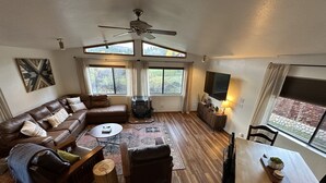 Amplia sala de estar con sofá de cuero marrón, una mesa de centro, una mesa de comedor con sillas, chimenea y ventanas grandes que ofrecen vistas a zonas verdes.
