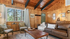 A cozy cabin interior with a wooden interior, a brown leather sofa, a wooden coffee table, and a view of the forest through a large window.