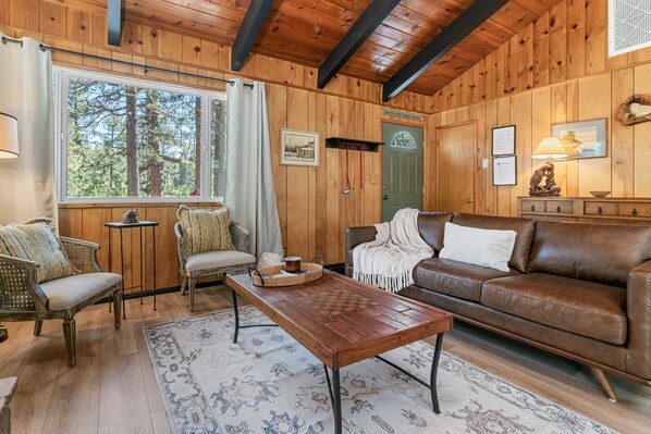 Ein gemütliches Blockhausschlafzimmer mit Holzausstattung, einem braunen Ledersofa, einem Holztisch und Blick auf den Wald durch ein großes Fenster.