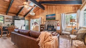 A cozy cabin interior with a wooden ceiling, a stone fireplace, a brown leather sofa, and a dining area with wooden chairs.