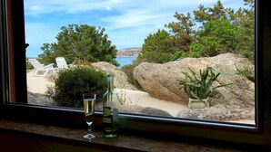 A view from a window overlooking a beach with lounge chairs, large rocks, and greenery. - Villa Mica in La Maddalena (La Maddalena)