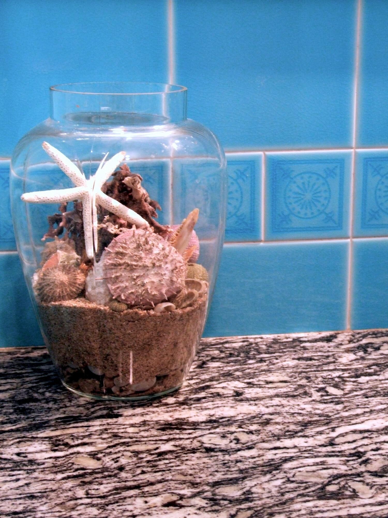 Un vase en verre transparent contenant une étoile de mer, un coquillage et d’autres éléments de décoration marine, posé sur un comptoir en granit.