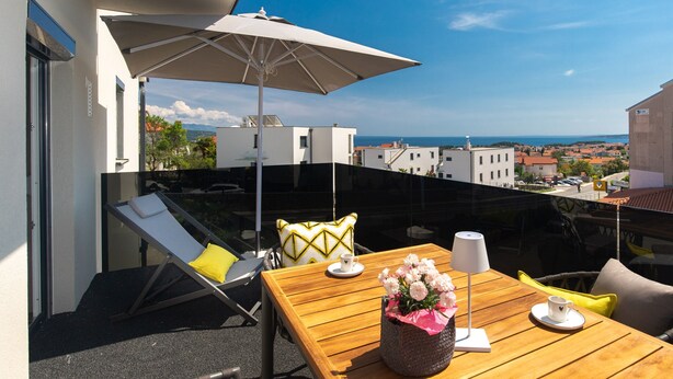 A balcony with a wooden table, a white umbrella, and a view of the sea and buildings. - Villa LuiSole apartment °2 with heated saltwater pool, family-friendly, sea view (Krk)