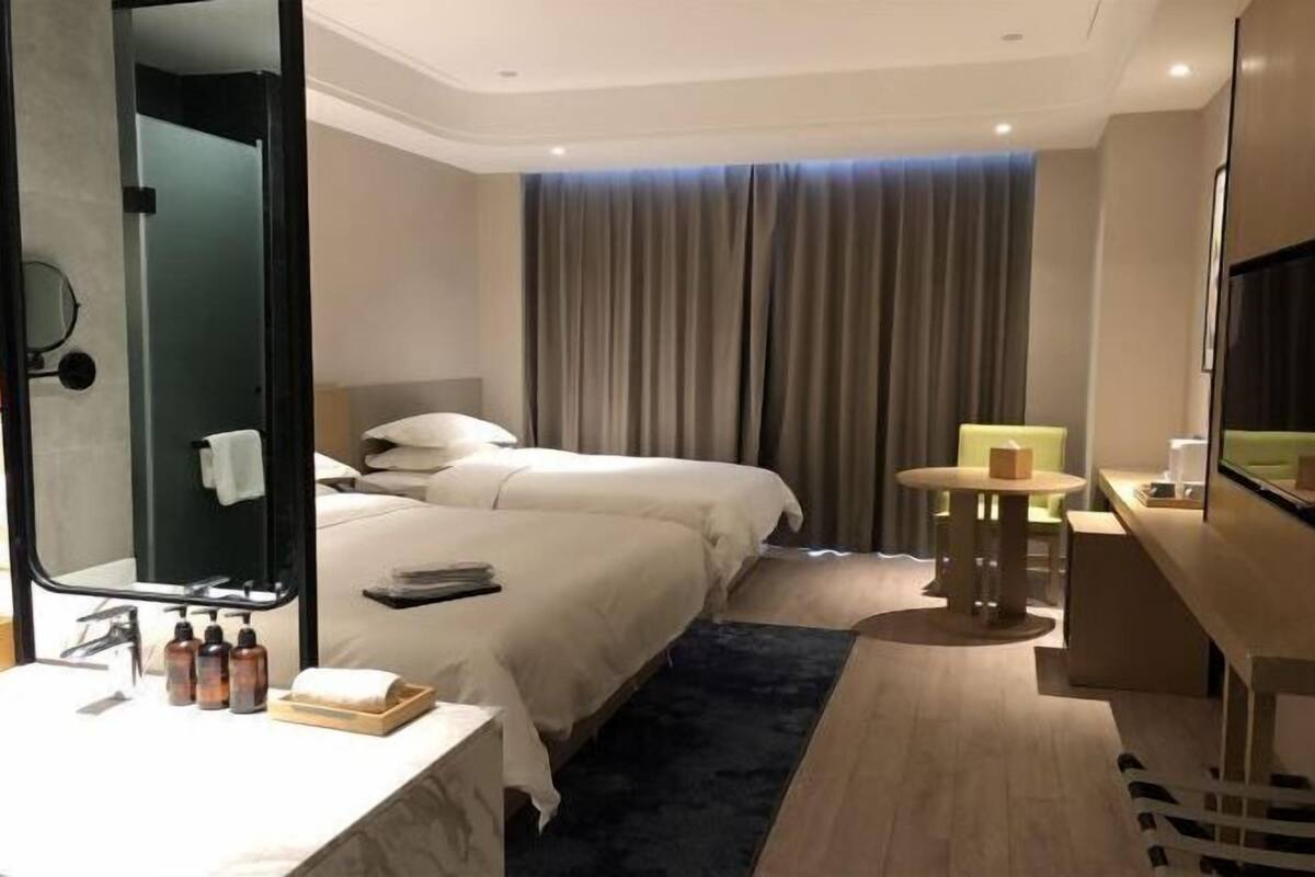 A hotel room with two beds, a desk, a mirror, a TV, and a bathroom with a sink and toiletries.